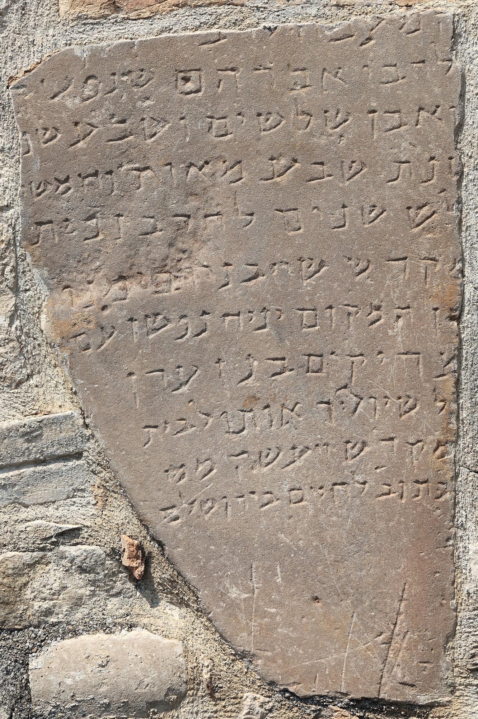 Photo of a weathered, irregular stone tablet, mounted into a brick wall, with a Hebrew inscription conforming to its shape.