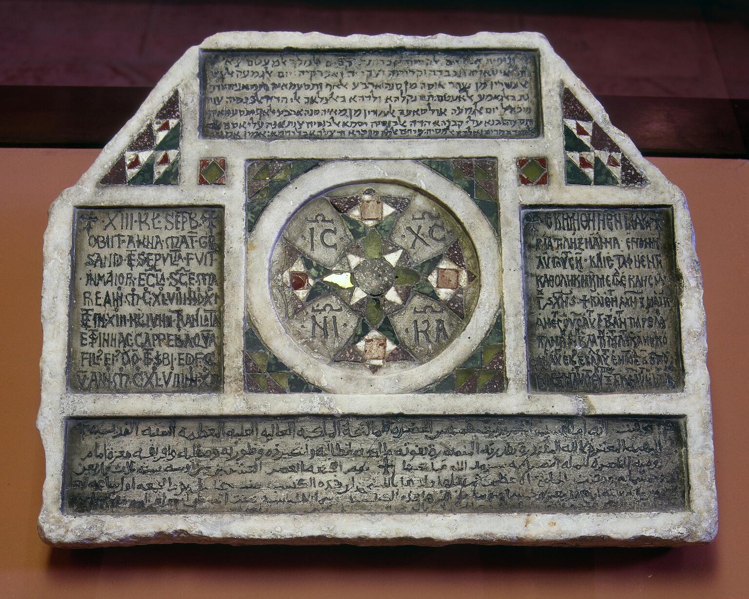 Photo of a hexagonal stone slab engraved with thickly-borded inscriptions in Latin, Hebrew, and Arabic, among which are geometric decorations, including, centrally, a circular pattern with four Latin letter pairs.