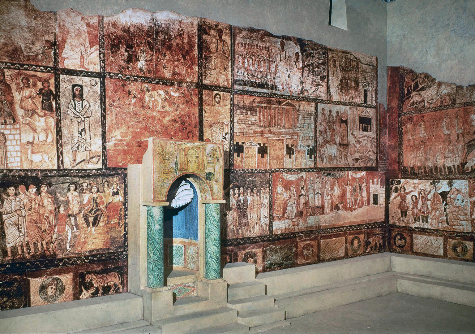 Colorful ancient wall paintings with biblical scenes and figures surround a small, elaborately decorated niche with stairs, set against a stone floor and walls in an archaeological restoration.