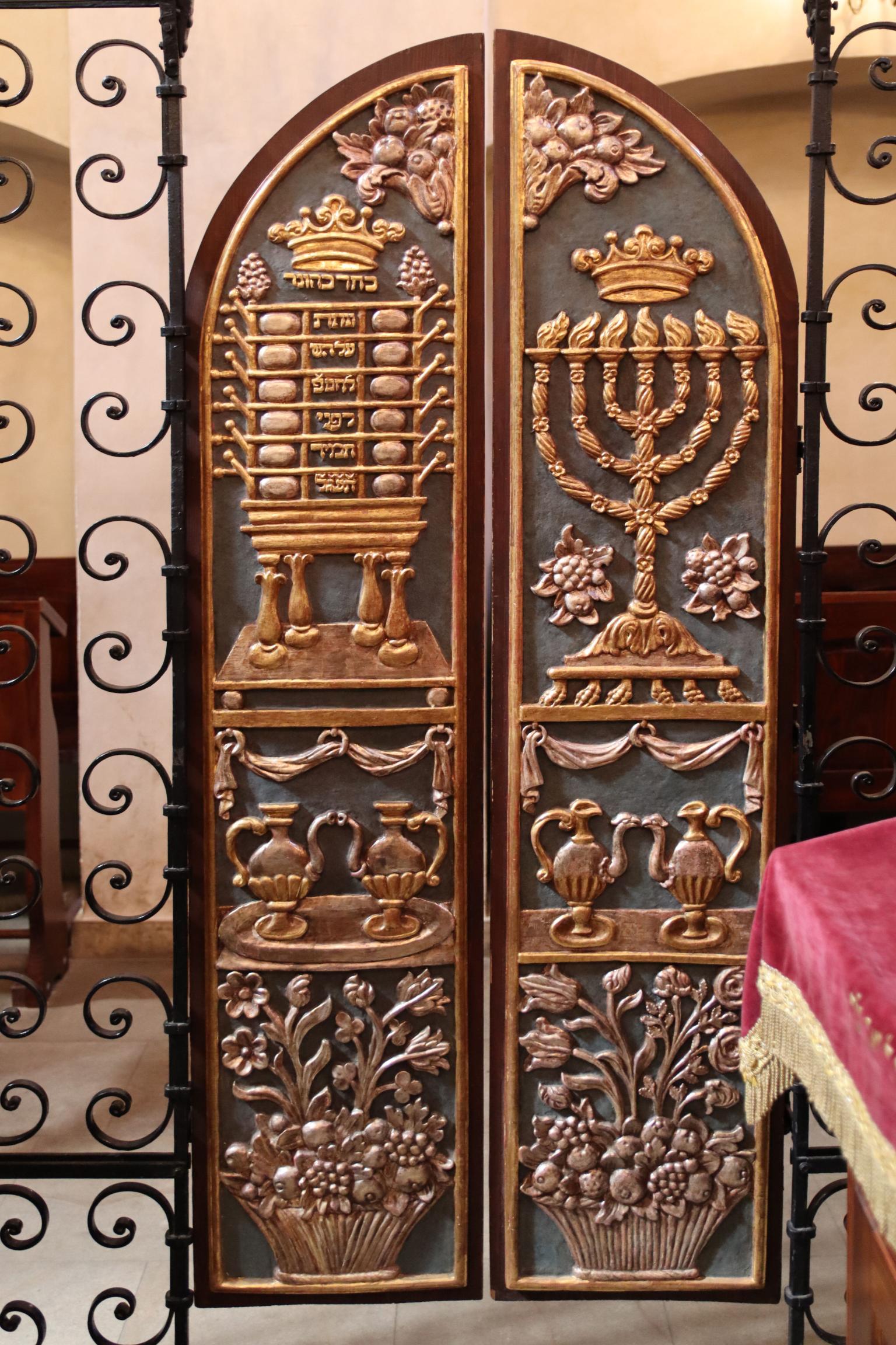 Set of curved doors with carving of candelabrum, peacock-shaped vessels, and basket of fruit and flowers on right side and table with compartments, peacock-shaped vessels, and basket of fruit and flowers on left side. 