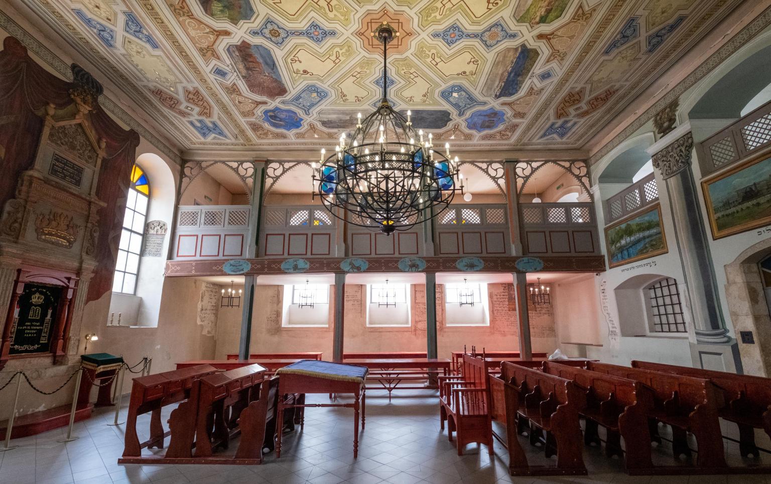 Photograph of interior room with second floor balcony, painted ceiling, rows of pews, and Torah ark on left wall. 