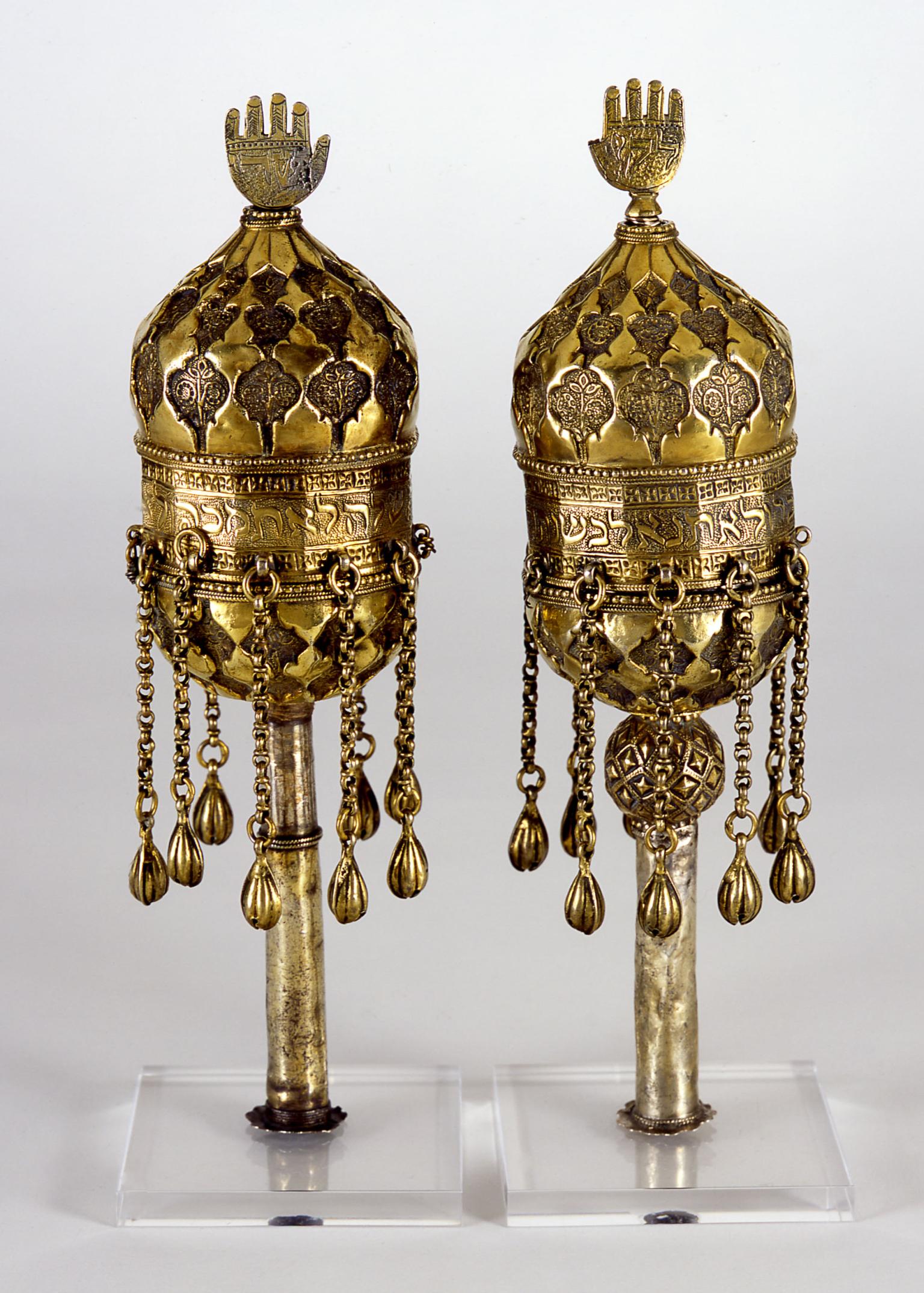 Two finials engraved with designs and Hebrew inscription, with chains hanging down from bottom and hand-shaped sculpture on top. 