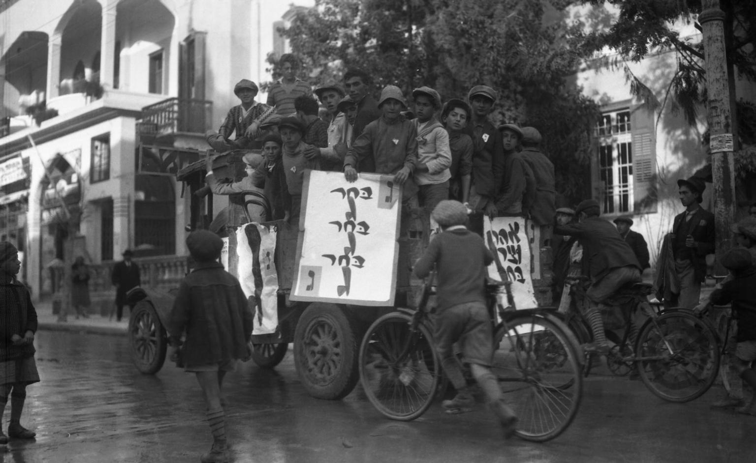 Photograph of men huddled together on a car holding billboards, surrounded by young men on bicycles following them down the street.