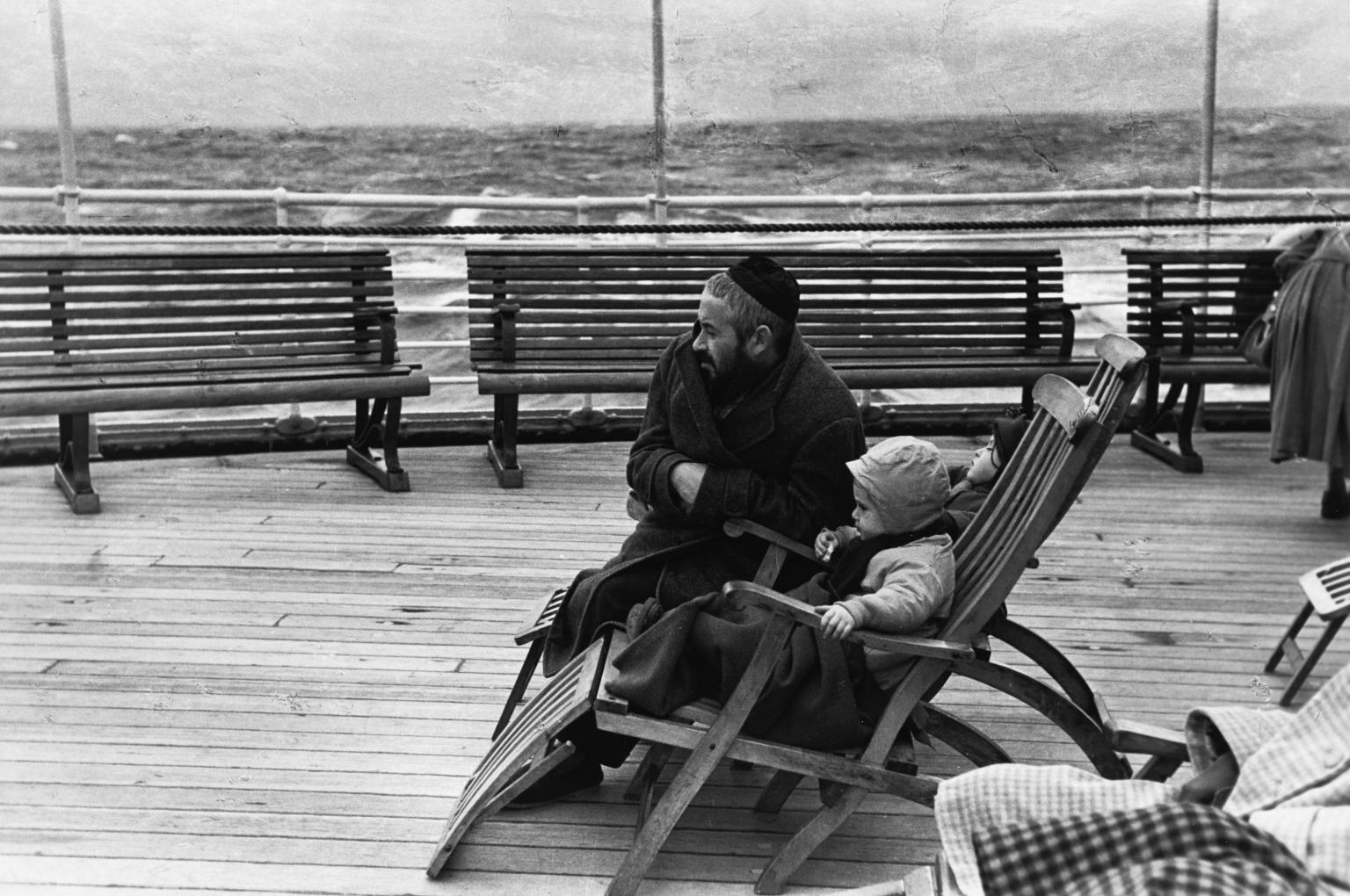 Photograph of man in a skullcap kneeling next to a child sitting in a wooden chair on boat deck, gazing into the distance to the left of the viewer.