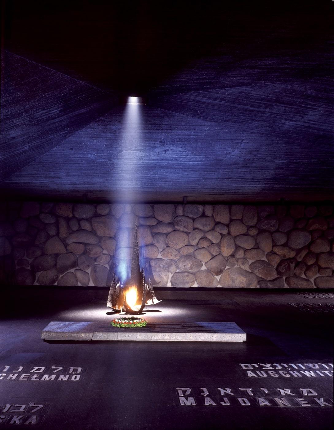 Memorial depicting flame burning from a base and illuminating a wall with an angular roof and basalt walls, with place names in English and Hebrew on the floor.