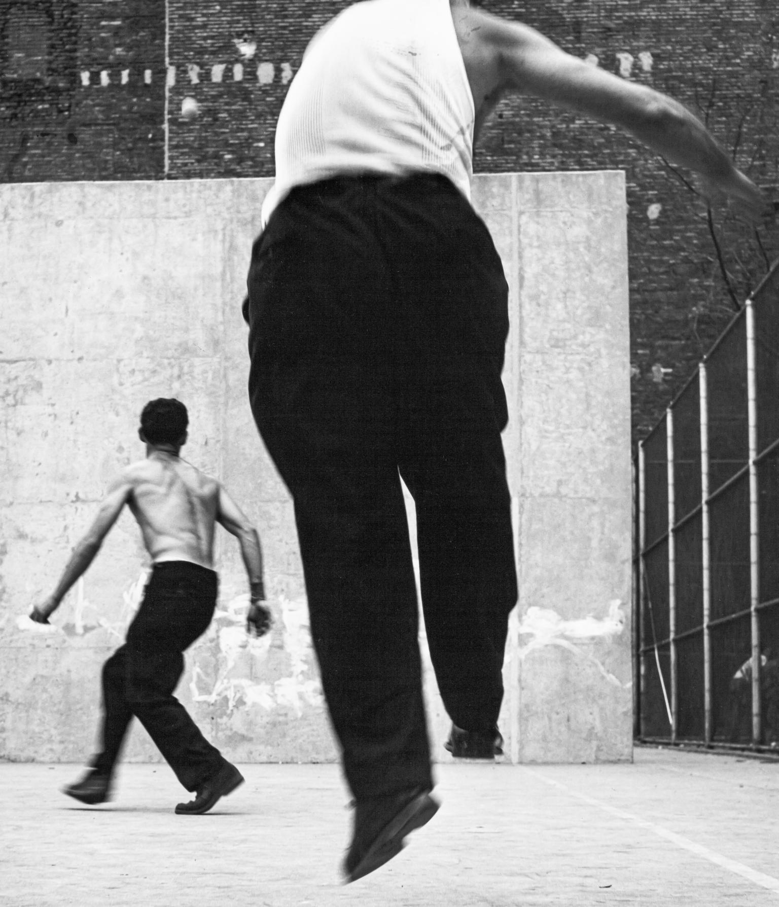 Photograph of two men playing handball, both of whom are turned away from the viewer and facing the wall. 