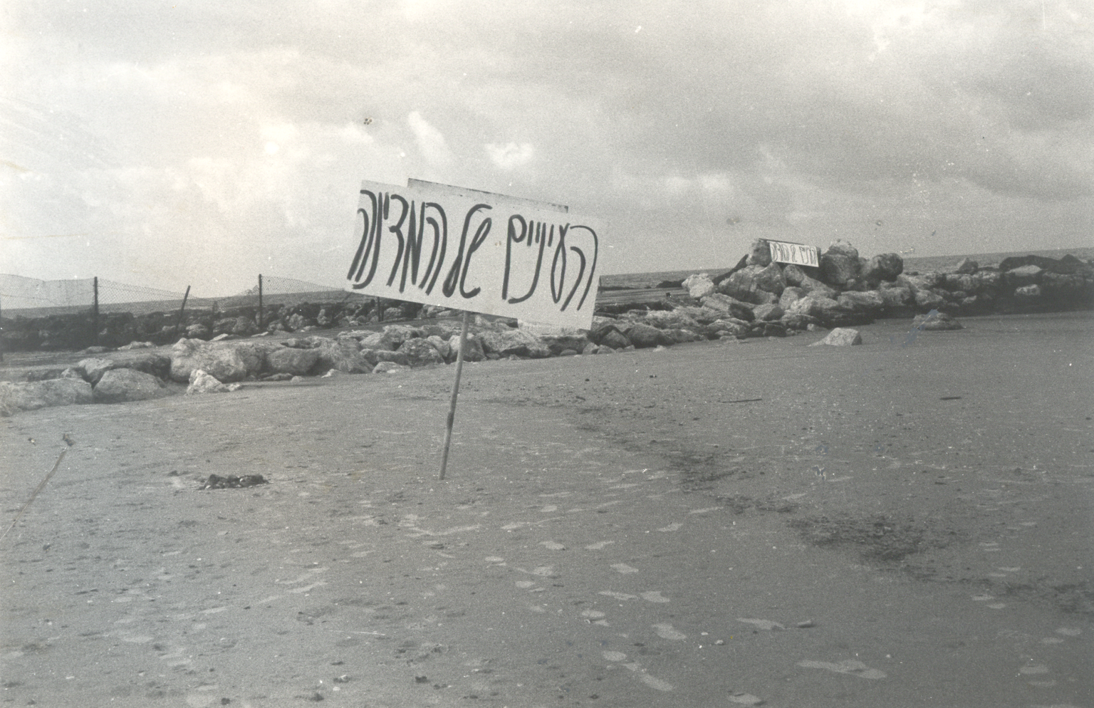 Photograph of Hebrew sign on empty beach with a rock jetty in background.