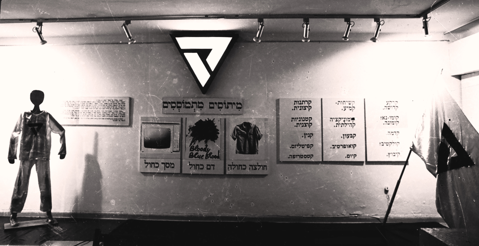 Photograph of wall with a mannequin to the left, posters with Hebrew text and an illuminated triangle on the wall, and a flag to the right.
