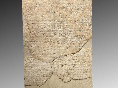 An ancient stone stela with faded, inscribed text in Greek, topped by a flower-like carving and featuring a pointed, decorated top. The stone shows visible cracks and repairs.