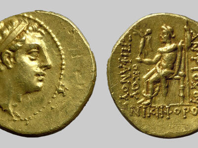Ancient gold coin. Obverse shows a right-facing profile with a diadem. Reverse depicts a seated figure holding an object, surrounded by inscriptions in Greek.