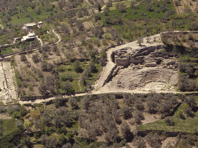 Aerial view of an ancient stone amphitheater surrounded by green fields, scattered trees, and ruins of other structures in a rural landscape.