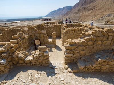 Ancient stone ruins with low walls and doorways under a clear sky, with mountains and the Dead Sea visible in the background. Tourists walk along a pathway nearby.