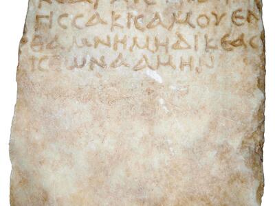 A weathered, rectangular stone slab featuring several lines of faded ancient Greek inscriptions carved into its surface.