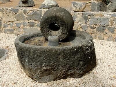 A large, round olive press consisting of a circular basin and wheel sits outdoors on gravel, surrounded by a low stone wall and other stone objects.