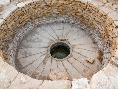 Circular stone basin with layered stone walls, a base with a radial pattern, and a round opening at the bottom. A few rocks are scattered inside.