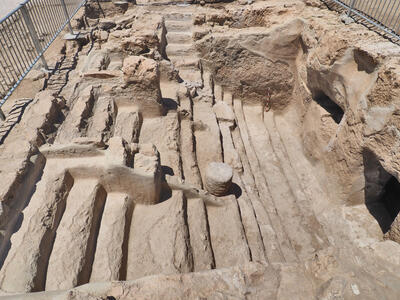 Ancient stone steps leading down into an area with vertical walls and some openings, surrounded by a metal fence. The area shows layered, weathered stone surfaces.