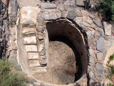 A round, stone-lined pit with built-in stone steps leading down to a dirt floor. The structure is surrounded by rocks and sparse vegetation.