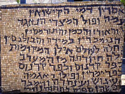 A rectangular stone mosaic with multiple lines of Hebrew script in dark tiles, set against a background of lighter-colored tiles. The mosaic appears weathered and is displayed outdoors.