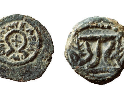 Two sides of an ancient, weathered coin: one side shows an x shape surrounded by an omega shape, encircled by Greek inscriptions, and the other features a tripod flanked by branches. The metal is rough and greenish.