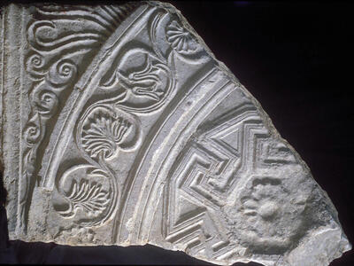 A fragment of carved stone features intricate floral and geometric patterns, including curved vines, leaf motifs, and repeating angular designs, with detailed, weathered textures visible on its surface.