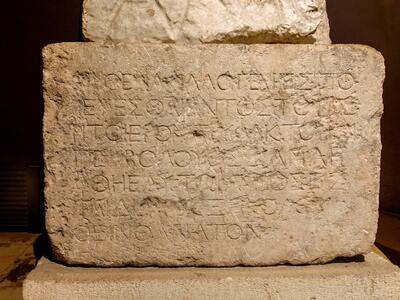 A rectangular ancient stone tablet with worn Greek inscriptions carved into its surface, displayed upright in a museum setting. The text is faded and partially eroded, making some letters difficult to read.
