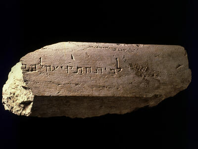 A weathered stone slab with an ancient Hebrew inscription carved into its surface, displayed against a black background. The stone is uneven and partially broken on the left side.