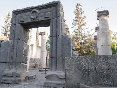 Ancient stone ruins with a large rectangular archway and several standing columns, surrounded by trees and greenery under a clear sky.