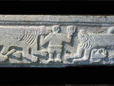 Ancient stone relief showing a human between two lions flanked by two eagles. A lion cub lying on its back nurses from the lion on the right.