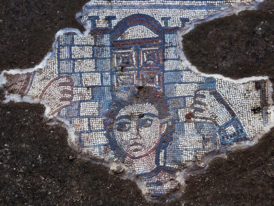 A fragment of an ancient mosaic depicts a human face and hands grasping an architectural structure with a gate in the center. The surrounding area shows dark, rough ground.
