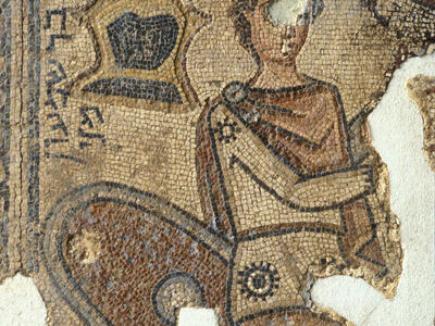 Ancient mosaic depicting a partially damaged figure in armor, with a helmet, shield, and sword in the background. Some areas of the mosaic are missing.