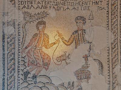 Ancient mosaic depicts two robed figures and a donkey. Greek text appears above the scene. There are areas of visible wear and missing tiles.