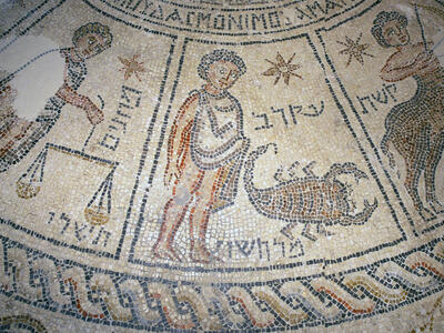 Ancient mosaic depicting three zodiac signs: Libra with scales, Scorpio with a scorpion, and Sagittarius as a centaur archer. Hebrew inscriptions and decorative patterns frame the figures.