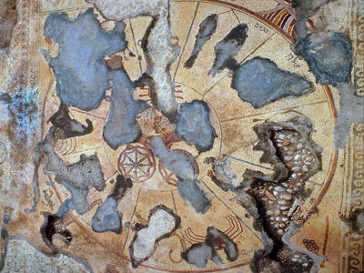 Ancient mosaic with significant damage and missing sections, featuring faded geometric patterns and portions of animals and human figures on a circular background. Some original intricate details are still visible around the edges.