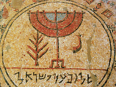 Ancient mosaic featuring a central menorah design, flanked by a plant and a shofar, with Hebrew text below, all set within a circular border amid intricate geometric patterns.