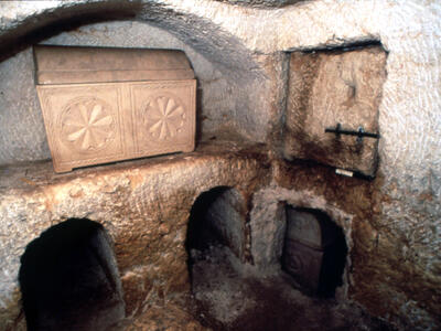Ancient stone tomb with arched niches and a decorated limestone ossuary carved with rosettes, set in a small, dimly lit rock-cut chamber.