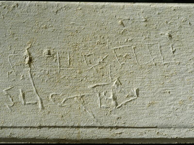 Graffiti scratched into a rough, light-colored stone surface, featuring uneven, hand-carved letters.