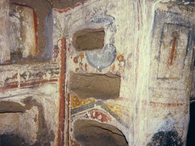 Ancient stone tomb with rectangular niches carved into the walls, decorated with faded frescoes and geometric patterns in red, yellow, and blue. Some niches appear empty; white mineral streaks run down the wall.
