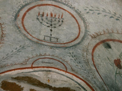 Ancient painting featuring a menorah with seven lit candles inside a bordered circle, surrounded by decorative patterns and arches on a stone surface.