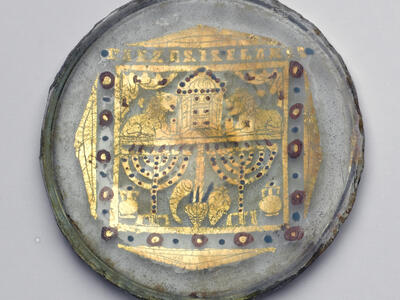 A round, ancient glass circle decorated with gold leaf showing two menorahs, ritual objects, and a central structure flanked by lions, with geometric and floral patterns.
