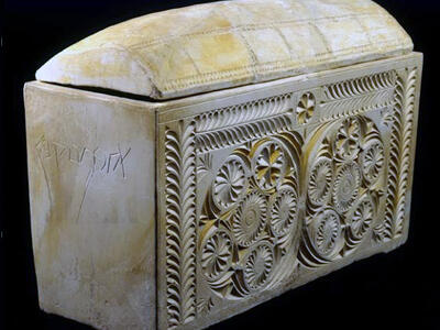 A rectangular carved limestone ossuary with intricate floral and geometric patterns on the front and a partially fitted lid. There is an inscription in an ancient script on the side.