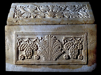 A stone ossuary with intricate carvings of grape clusters and leaves, a central palm tree motif, and an inscription in ancient script above the central panel.