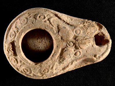An ancient clay oil lamp with a large central opening and a smaller opening near the spout, set against a black textured background. The lamp is oval shaped with decorative patterns around its surface.