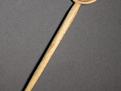 Bone object shaped like a small disk with a long, straight handle. The disk features three spiral patterns arranged in a triangular formation.