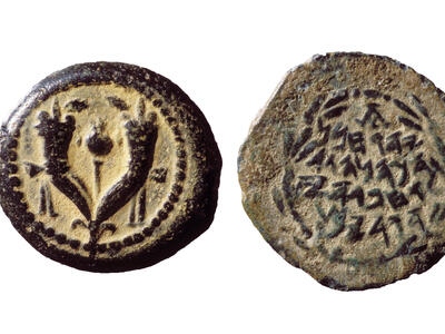 Two sides of an ancient coin: one side shows two joined cornucopias with a pomegranate in between, and the other side has an inscription in Paleo-Hebrew surrounded by a laurel wreath. The coin is aged and worn.