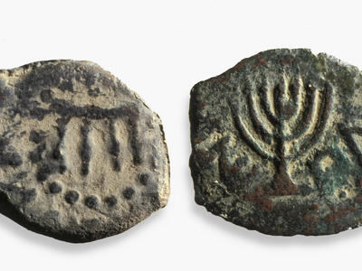 Two sides of an ancient bronze coin: one shows a four-legged table surrounded by dots, and the other features a seven-branched menorah. Both sides have faint inscriptions. The coin appears worn and weathered.