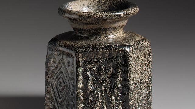 Photo of a dark glass vase with a wide, flared rim and hexagonal base, featuring geometric patterns.