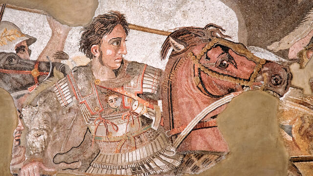 Ancient mosaic depicting a warrior in ornate armor, holding a spear, riding a horse into battle. The warrior has wavy hair and a determined expression and is surrounded by other soldiers. The background is partially damaged.