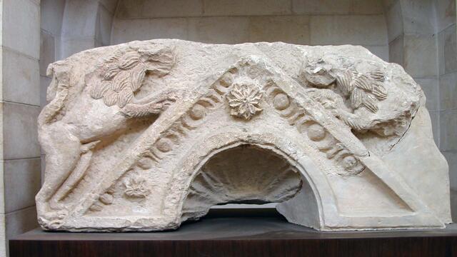 A weathered stone fragment featuring two carved lions surrounding a triangular roof with an arched opening.