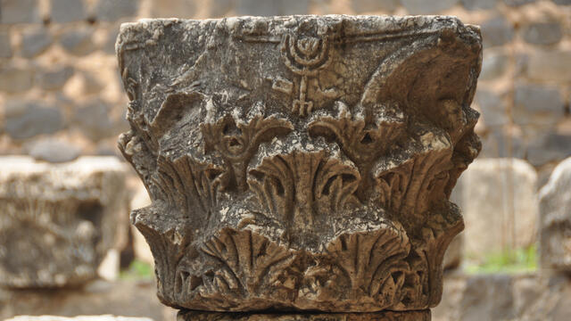 A weathered stone capital featuring carved vegetal motifs and a prominent menorah symbol on top, situated outdoors against a background of rough stone blocks.