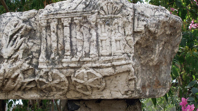 A weathered stone block with a carved relief depicting a chariot with columns and an arched door, set outdoors with foliage and flowers in the background.